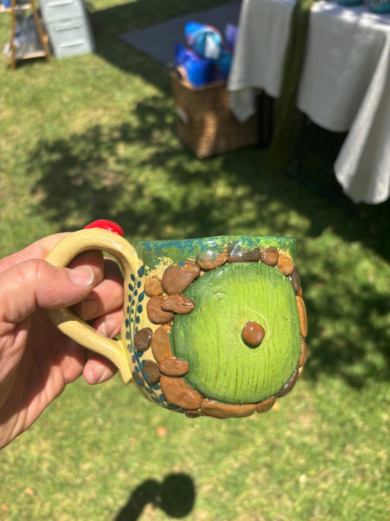 May include: A ceramic mug featuring a sculpted green circular door design surrounded by brown stone-like textures. The mug has a light tan handle and a green rim, with small blue leaf patterns painted along the side near the handle attachment.