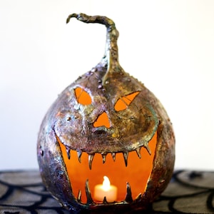 May include: A metallic, textured pumpkin with a carved jack-o'-lantern face and a lit candle inside. The pumpkin is sitting on a black spiderweb patterned surface.