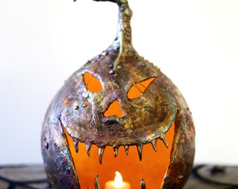 Könnte beinhalten: Ein metallischer, strukturierter Kürbis mit einem geschnitzten Jack-o'-Lantern-Gesicht und einer brennenden Kerze im Inneren. Der Kürbis steht auf einer schwarzen Oberfläche mit Spinnennetz-Muster.
