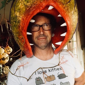 K&ouml;nnte beinhalten: Eine Person tr&auml;gt ein wei&szlig;es T-Shirt mit dem Text "Ich liebe K&auml;tzchen" und einem Cartoon-Katzenmotiv. Die Person tr&auml;gt ein gro&szlig;es, rot-gr&uuml;nes, pflanzenartiges Kost&uuml;m mit einem gro&szlig;en Mund.