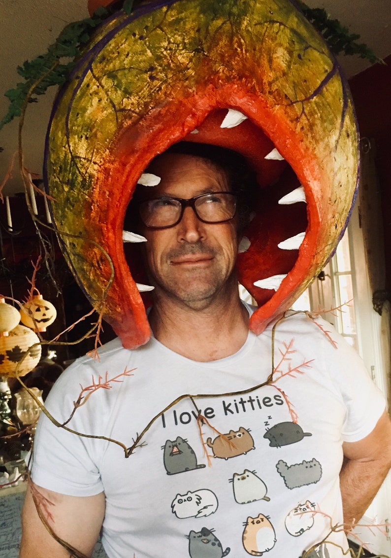 K&ouml;nnte beinhalten: Eine Person tr&auml;gt ein wei&szlig;es T-Shirt mit dem Text "Ich liebe K&auml;tzchen" und einem Cartoon-Katzenmotiv. Die Person tr&auml;gt ein gro&szlig;es, rot-gr&uuml;nes, pflanzenartiges Kost&uuml;m mit einem gro&szlig;en Mund.