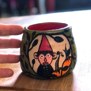 May include: Hand-painted ceramic mug featuring a cartoon character with a red hat and a surprised expression. The mug has a red interior and is decorated with green foliage and an orange pumpkin. The mug is on a wooden surface.