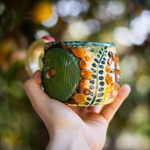 May include: A ceramic mug featuring a raised, circular green door design with a central knob, surrounded by textured orange and brown stones, green leaf patterns, and small red and white mushroom accents on a cream-colored glazed surface.
