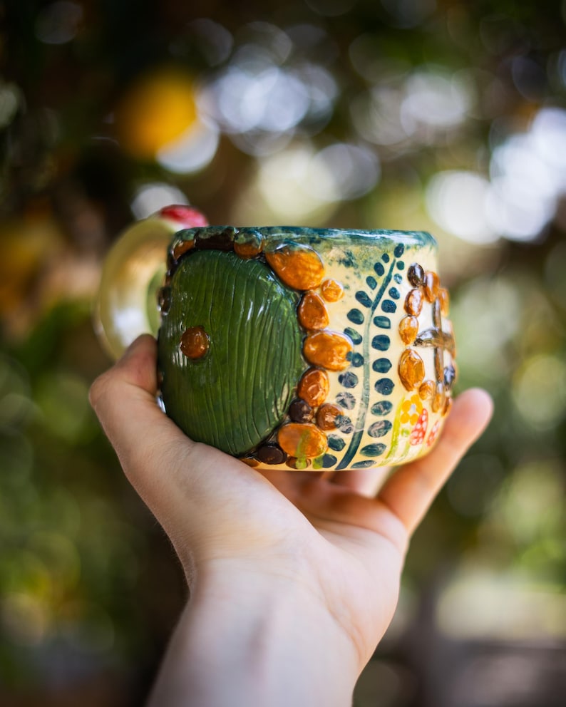 May include: A ceramic mug featuring a raised, circular green door design with a central knob, surrounded by textured orange and brown stones, green leaf patterns, and small red and white mushroom accents on a cream-colored glazed surface.