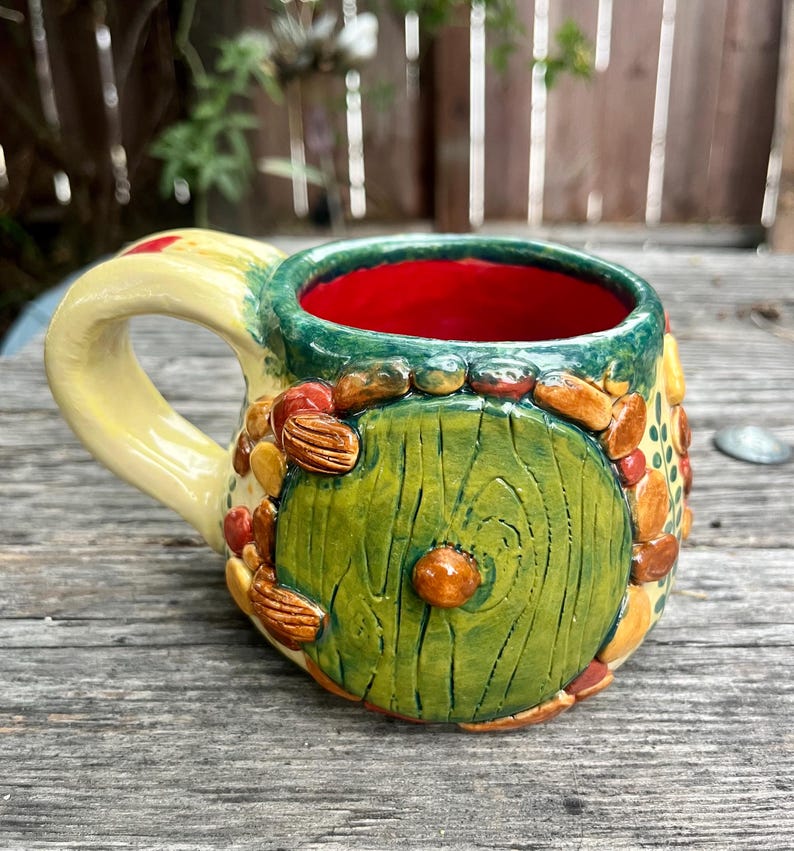 May include: A ceramic mug featuring a sculpted green circular door design on the front, surrounded by small brown and orange pebble-like accents. The mug has a glossy yellow handle, a vibrant red interior, and a green rim with painted leaf details.
