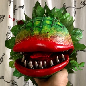 May include: A handmade puppet of a carnivorous plant with a large red mouth, sharp teeth, and a green body. The puppet is surrounded by green leaves.