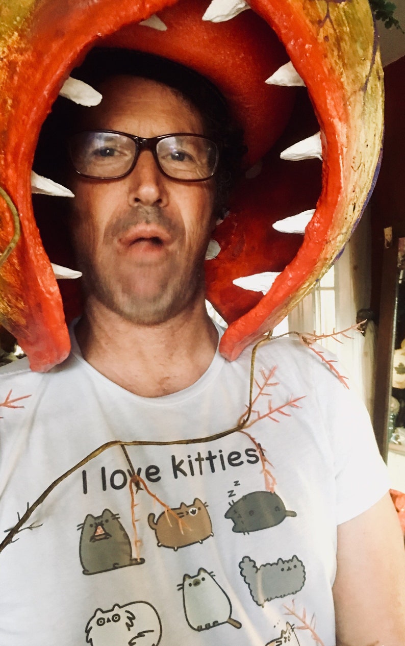 K&ouml;nnte beinhalten: Eine Person tr&auml;gt ein rotes und wei&szlig;es Kost&uuml;m einer fleischfressenden Pflanze mit einem wei&szlig;en T-Shirt, auf dem "Ich liebe K&auml;tzchen" steht und ein Motiv mit mehreren Comic-Katzen.
