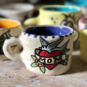 May include: A white ceramic mug with a blue rim and a blue and black bird tattoo design. The tattoo includes a red heart with the word "MOM" on a banner.