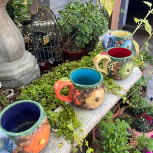 May include: Three ceramic mugs featuring a round door design on the exterior. The mugs have vibrant interior colors including blue, red, and purple, with textured, earthy-toned exteriors decorated with small sculpted leaves and circular door motifs.