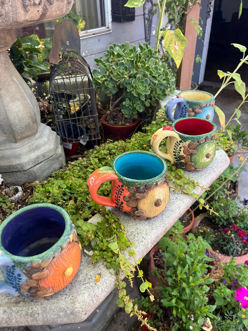 May include: Three ceramic mugs featuring a round door design on the exterior. The mugs have vibrant interior colors including blue, red, and purple, with textured, earthy-toned exteriors decorated with small sculpted leaves and circular door motifs.
