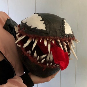 Puede incluir: Una máscara de Venom de papel maché en blanco y negro con una lengua de fieltro roja. La máscara tiene una boca detallada con dientes afilados y un ojo blanco.