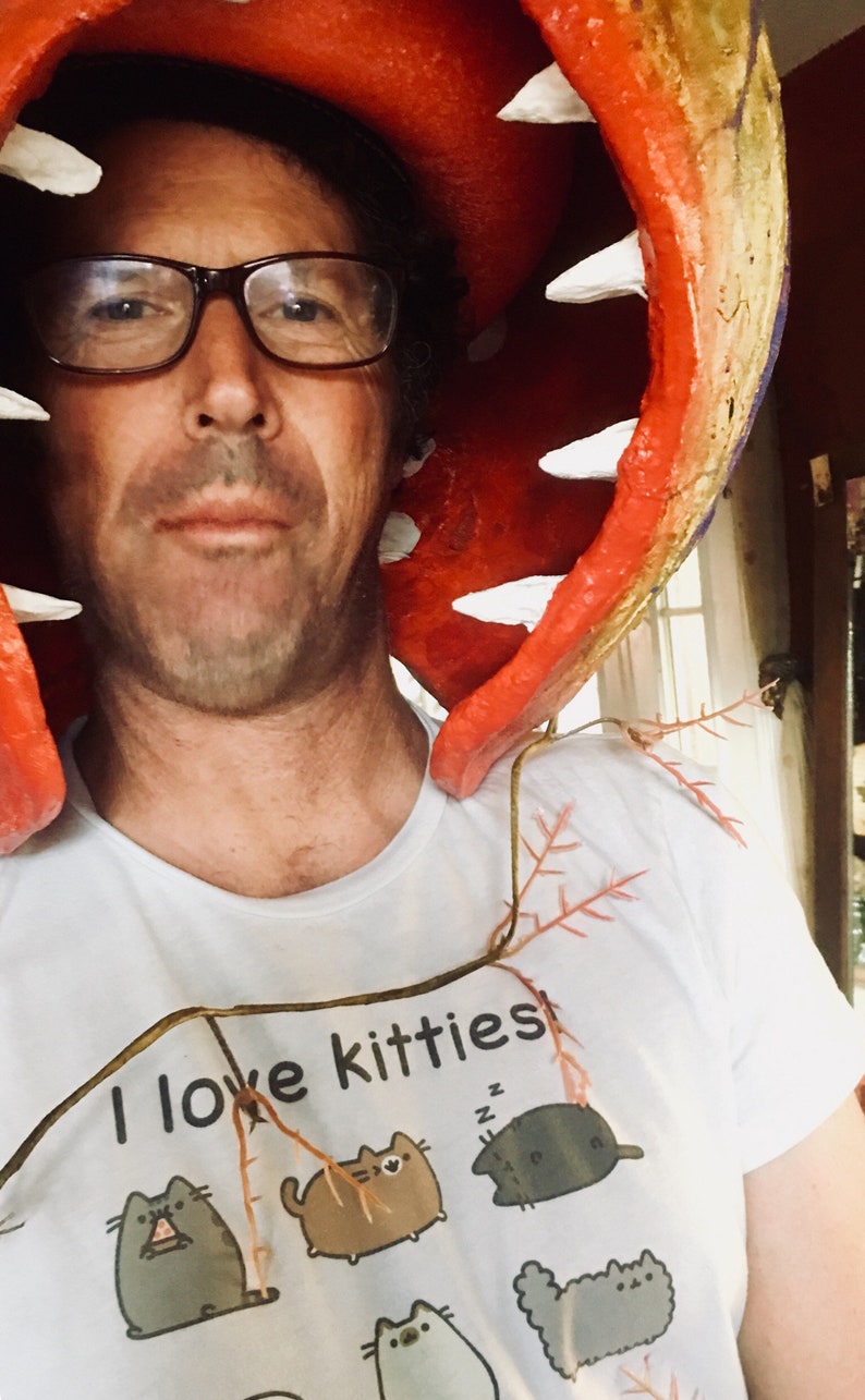 K&ouml;nnte beinhalten: Eine Person tr&auml;gt ein wei&szlig;es T-Shirt mit dem Text "I love kitties" und einem Cartoon-Katzenmotiv. Die Person tr&auml;gt ein rot-gr&uuml;nes, pflanzenartiges Kost&uuml;m mit wei&szlig;en Z&auml;hnen.