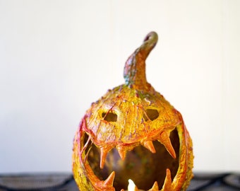 May include: A Halloween pumpkin carved with a scary face and lit from inside with a candle. The pumpkin is painted in shades of orange, yellow, and red, and has a textured surface. It is sitting on a black spiderweb tablecloth.