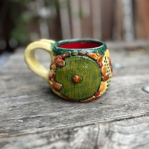 May include: A ceramic mug featuring a sculpted green circular door with a central knob, surrounded by textured brown and orange stones. The mug has a pale yellow handle, a dark green rim, and a vibrant red interior, with painted green leaf details.
