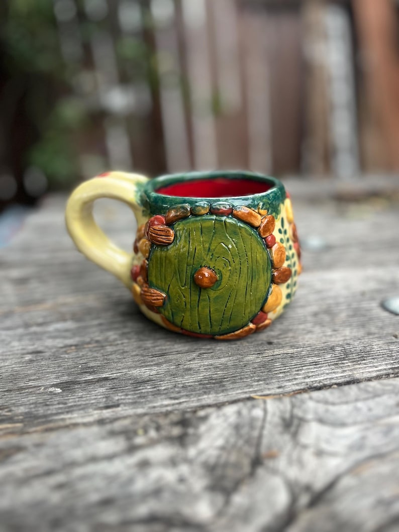 May include: A ceramic mug featuring a sculpted green circular door with a central knob, surrounded by textured brown and orange stones. The mug has a pale yellow handle, a dark green rim, and a vibrant red interior, with painted green leaf details.