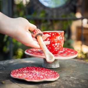 May include: A whimsical red and white polka-dotted ceramic teacup, saucer, and spoon set. The spoon is shaped like a mushroom. The teacup and saucer sit on a small pedestal. A second saucer is in the foreground.