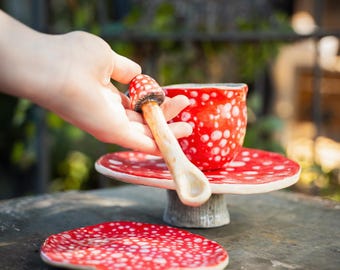 May include: A whimsical red and white polka-dotted ceramic teacup, saucer, and spoon set. The spoon is shaped like a mushroom. The teacup and saucer sit on a small pedestal. A second saucer is in the foreground.