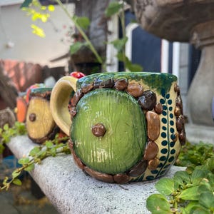 May include: A ceramic mug featuring a round green door design on the front, surrounded by sculpted brown stones and a light yellow handle. The mug has a textured surface with dark green accents and a vertical pattern of small dark green dots.