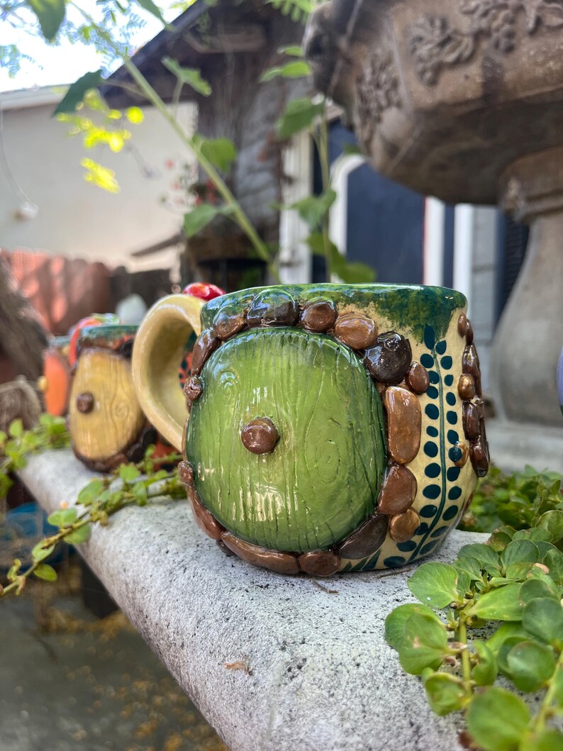 May include: A ceramic mug featuring a round green door design on the front, surrounded by sculpted brown stones and a light yellow handle. The mug has a textured surface with dark green accents and a vertical pattern of small dark green dots.