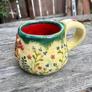 May include: A ceramic mug with a pale yellow exterior featuring hand-painted blue and orange floral patterns and a raised textured cluster of brown and tan circular shapes. The mug has a bright red interior and a dark green rim with a thick handle.