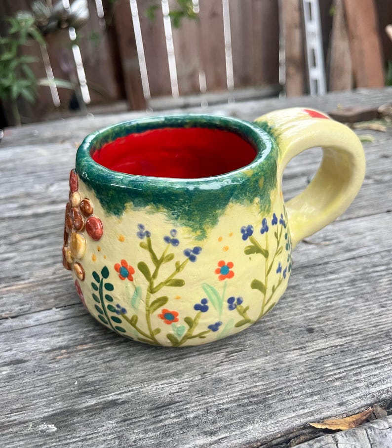 May include: A ceramic mug with a pale yellow exterior featuring hand-painted blue and orange floral patterns and a raised textured cluster of brown and tan circular shapes. The mug has a bright red interior and a dark green rim with a thick handle.