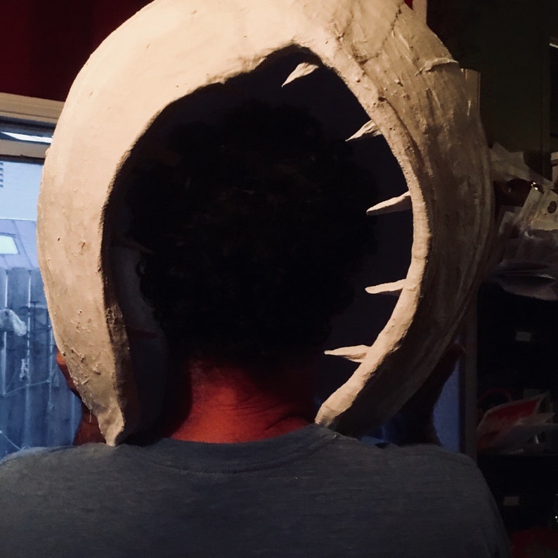 K&ouml;nnte beinhalten: Eine wei&szlig;e Gipsmaske mit einem gro&szlig;en, offenen Mund und scharfen Z&auml;hnen. Die Maske wird hinter dem Kopf einer Person gehalten.