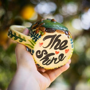 May include: A ceramic mug with a cream-colored base featuring the hand-painted black text The Shire and small red hearts. The top of the mug is sculpted to resemble a green hill with brown stones, and the handle is decorated with green leaves.
