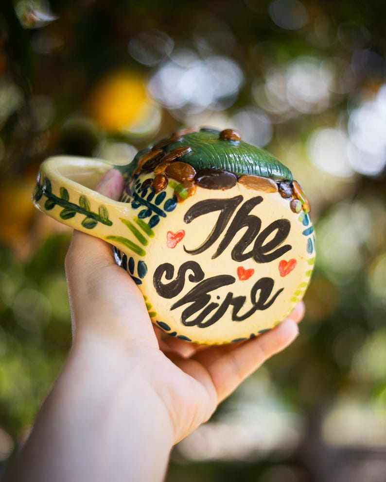 May include: A ceramic mug with a cream-colored base featuring the hand-painted black text The Shire and small red hearts. The top of the mug is sculpted to resemble a green hill with brown stones, and the handle is decorated with green leaves.