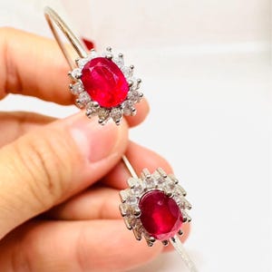 Puede incluir: Un brazalete abierto de tono plateado con dos piedras preciosas ovaladas de color rojo vibrante, cada una rodeada por un halo de pequeñas piedras claras y brillantes. La banda de metal tiene un acabado texturizado y un diseño simétrico.