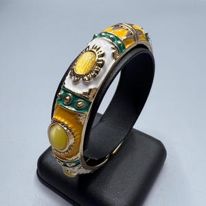 Może przedstawiać: Otwierana bransoleta typu bangle z podstawą z metalu w kolorze złota oraz sekcjami z białej, żółtej i turkusowej emalii. Bransoleta jest ozdobiona owalnymi żółtymi kamieniami typu kaboszon oraz małymi złotymi nitami wkomponowanymi w emalię.