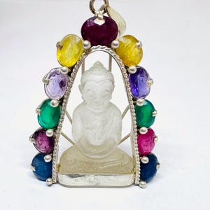 May include: A pendant featuring a carved, translucent, clear stone Buddha figure seated in a meditative pose. The figure is framed by a silver arch adorned with ten faceted, multi-colored gemstones, including yellow, purple, green, pink, and blue.