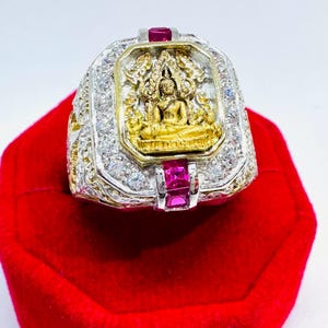 May include: A silver-toned ring featuring a central gold-coloured relief of a seated figure in a meditative pose. The rectangular setting is bordered by clear white stones and accented with two vibrant red rectangular stones at the top and bottom.