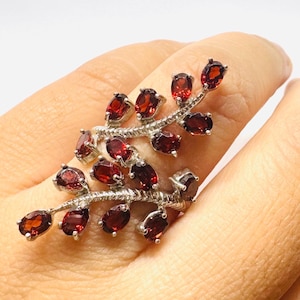Peut inclure: Une bague en argent au design en forme de branche, ornée de multiples pierres précieuses grenat rouge foncé de taille ovale. L'anneau en métal présente un motif texturé en chevrons imitant une tige, avec les pierres serties en feuilles.