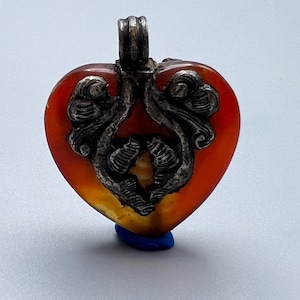 May include: A heart-shaped pendant made of translucent orange stone featuring an ornate, dark metallic overlay with swirling, leaf-like patterns. The pendant includes a sturdy metal bail at the top for attachment, showcasing a contrast between the smooth, polished surface of the stone and the textured, darkened metal detailing.
