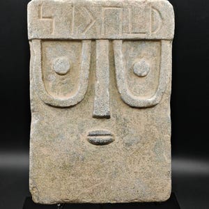May include: A rectangular gray stone slab featuring a carved face with large U-shaped eyes, a central nose, and a small mouth. The top section contains a horizontal band with engraved geometric symbols that resemble letters or ancient markings.