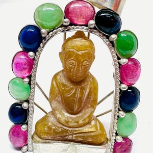 Puede incluir: Un colgante de plata con una figura de Buda tallada en piedra amarilla en el centro, encerrada en un marco arqueado. El marco está decorado con una serie de piedras preciosas cabujón redondas y pulidas en tonos vivos de rosa, verde, azul y negro.