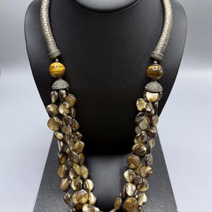 Può includere: Una collana a più fili con dischi di conchiglia marroni iridescenti e due perline rotonde di occhio di tigre color marrone dorato. La collana presenta accenti in metallo argentato testurizzato e un elemento rigido e curvo nella parte superiore.
