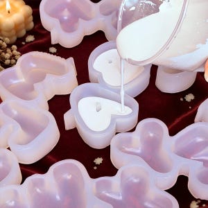 Pode incluir: Um conjunto de moldes de silicone translúcidos com formas variadas, incluindo corações e flores, com cera líquida branca a ser vertida de um jarro de plástico transparente para um molde em forma de coração com um pavio central.