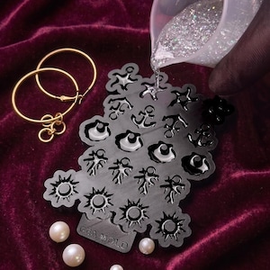May include: A black silicone mold with various shapes including anchors, suns, and shells, labeled GEA MOLD. A clear liquid with silver glitter is being poured into the mold. Two gold hoop earrings and three white pearls are placed nearby.