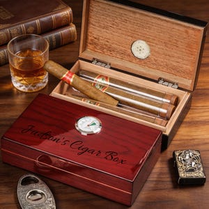 以下が含まれることがあります： 蓋にJudson's Cigar Boxの文字が刻印された、光沢のあるダークレッドの木製シガーボックス。アナログ湿度計が内蔵されています。中には透明なチューブに入った葉巻、琥珀色の液体が入ったグラス、金属製のシガーカッター、ライターが収められています。