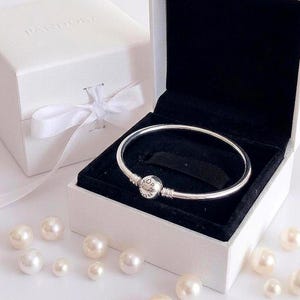 Puede incluir: Una pulsera rígida de plata con un cierre redondo que muestra el logotipo de Pandora, situada dentro de un joyero blanco abierto con interior de terciopelo negro. Una caja de regalo blanca cerrada con lazo y varias perlas blancas están cerca.