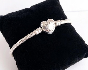 May include: A silver snake chain bracelet featuring a heart-shaped clasp. The clasp is engraved with the word PANDORA in a circular pattern. The bracelet has a smooth, metallic finish and is designed to hold charms along its flexible chain structure.