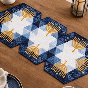 May include: A quilted table runner featuring a repeating geometric pattern in shades of navy and light blue. The design includes white star shapes and embroidered gold menorahs. The fabric has a textured, stitched finish with floral accents.