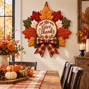 Puede incluir: Un tapiz de tela acolchada con forma de corona de hojas otoñales en tonos rojo, naranja, amarillo y verde. El centro presenta un panel circular con el texto Give Thanks, acentuado por un lazo de tela a cuadros en la parte inferior.