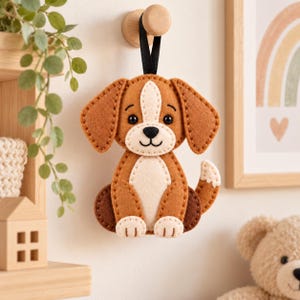 May include: A brown and cream felt dog ornament with floppy ears, a black stitched nose, and a small tail. The dog is suspended by a black ribbon from a wooden wall hook, featuring visible blanket stitching along the edges of the felt pieces.