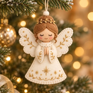 Pode incluir: Um enfeite de anjo em feltro com vestido e asas na cor creme, decorado com padrões de folhas bordadas em dourado e uma estrela central. A figura tem cabelos castanhos com uma tiara dourada, olhos fechados e mãos juntas em posição de oração.