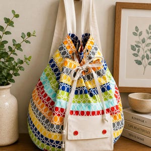 Op de afbeelding: Een rugzak met trekkoord voorzien van een levendig patroon van kleurrijke cirkels in geel, oranje, rood, blauw en groen. De tas heeft off-white canvas schouderbanden en een bijpassend voorvak dat sluit met twee kleine rode knopen en een koord.