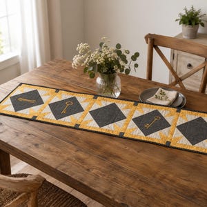 May include: A rectangular quilted table runner featuring a repeating pattern of dark gray diamond shapes centered within yellow and white starburst blocks. Each dark gray diamond contains a gold-colored embroidered key motif.