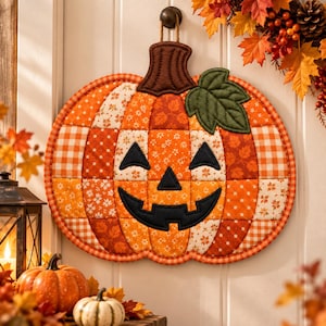May include: A quilted fabric pumpkin wall hanging featuring a patchwork design of orange, white, and brown autumn-themed patterns. The pumpkin has a dark brown stem, a green leaf, and a black stitched jack-o'-lantern face with a wide grin.