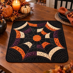 Peut inclure: Un set de table matelassé carré présentant un cercle orange central entouré d'un motif de toile d'araignée rayonnant. Le design intègre des segments incurvés en tissus noir, orange et crème avec des motifs floraux et étoilés.